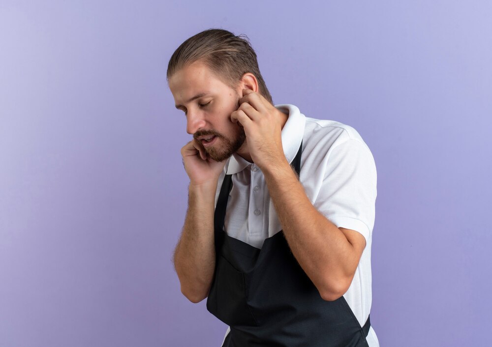 Unpleased young handsome barber wearing uniform putting fingers in ears with closed eyes isolated on purple background with copy space